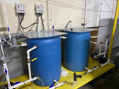 Barrels containing coolant are connected to a plumbing system that pipes around the facility at ACR Machine Co. Also visible is a box-like parts tumbling machine. 