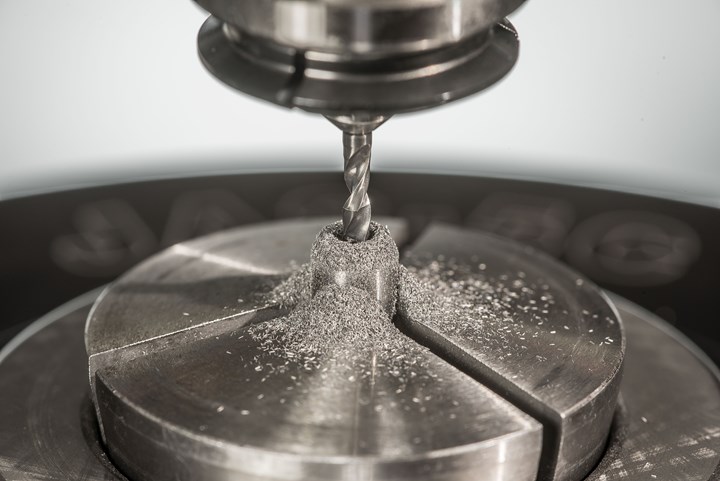 Dust-like piles of chips surround a fluted tool boring into a tungsten blank held in a chuck on the table of a CNC machining center.