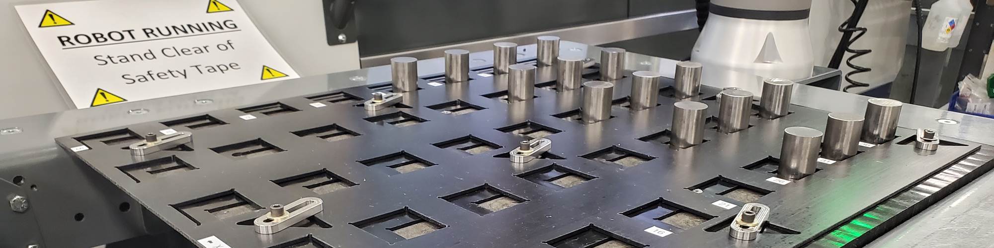 A system of overlapping trays features adjustable square openings that stage multiple, metal workpieces for pickup and loading into a CNC machine tool with a robot arm. 