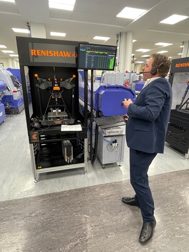 Guy Brown, development manager of Renishaw Central, shows off an Equator gaging system while dressed in a blue suit, standing on the Renishaw shop floor in the United Kingdom