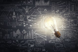 lightbulb that is on in front of a background of a black chalkboard with various business notes and drawing on it