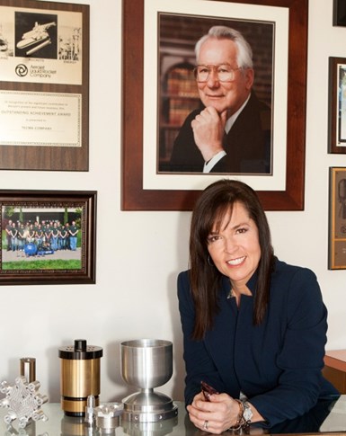 A photo of TECMA president Sonia Susac sitting beneath a portrait of her father, founder Fred Schwarz