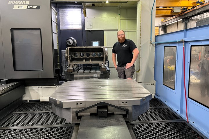 A photo of a man standing on the loading area of a Doosan HM 1250W horizontal machining center. A part is to one side of him, while a pallet is in front.