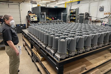 A photo of MTH Pumps owner Tim Tremain standing next to a large collection of Freon pumps. A robotic welding cell rises from the back of the room.