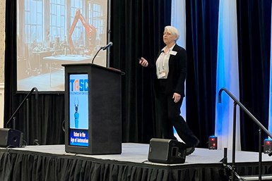 Andra Keay onstage at TASC - The Automated Shop Conference. Her presentation plays in the background, with a photo of a robotic arm onscreen.