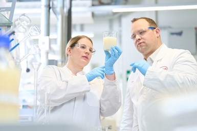 Photo of 2 people in a lab setting looking at a lab container full of opaque cream liquid one is holding.