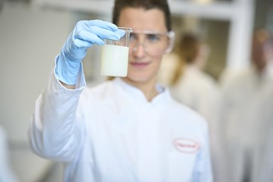 Image of woman blurred in background wearing lab coat and holding lab beaker with cloudy white substance in it