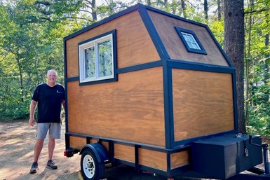 Photo of Tim Chicoine standing in the woods next to his custom-built, trailer mounted camper, the  
