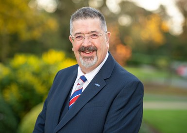 Photo of BVAA's Rob Barlett standing outside in front of a row of bushes. Rob has glasses and a graying short beard and is wearing a navy blue suit and red, white and blue tie.
