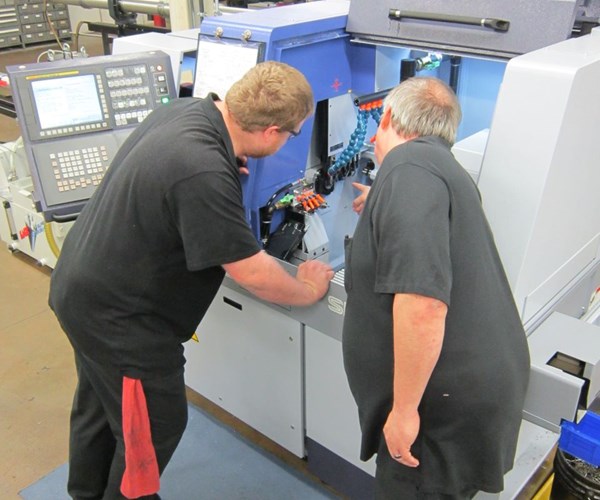 a baby boomer and a younger employee stand in front of a machine discussing a manufacturing process